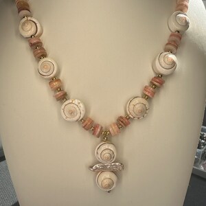 May include: A white and pink seashell necklace with a gold accent. The necklace features a pendant with three seashells.