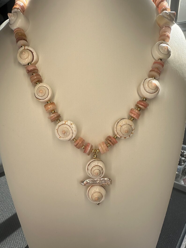 May include: A white and pink seashell necklace with a gold accent. The necklace features a pendant with three seashells.