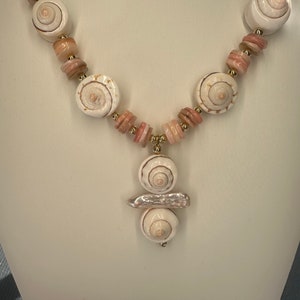 May include: A white and pink shell necklace with a pendant made of three shells. The pendant is attached to the necklace with a gold chain.