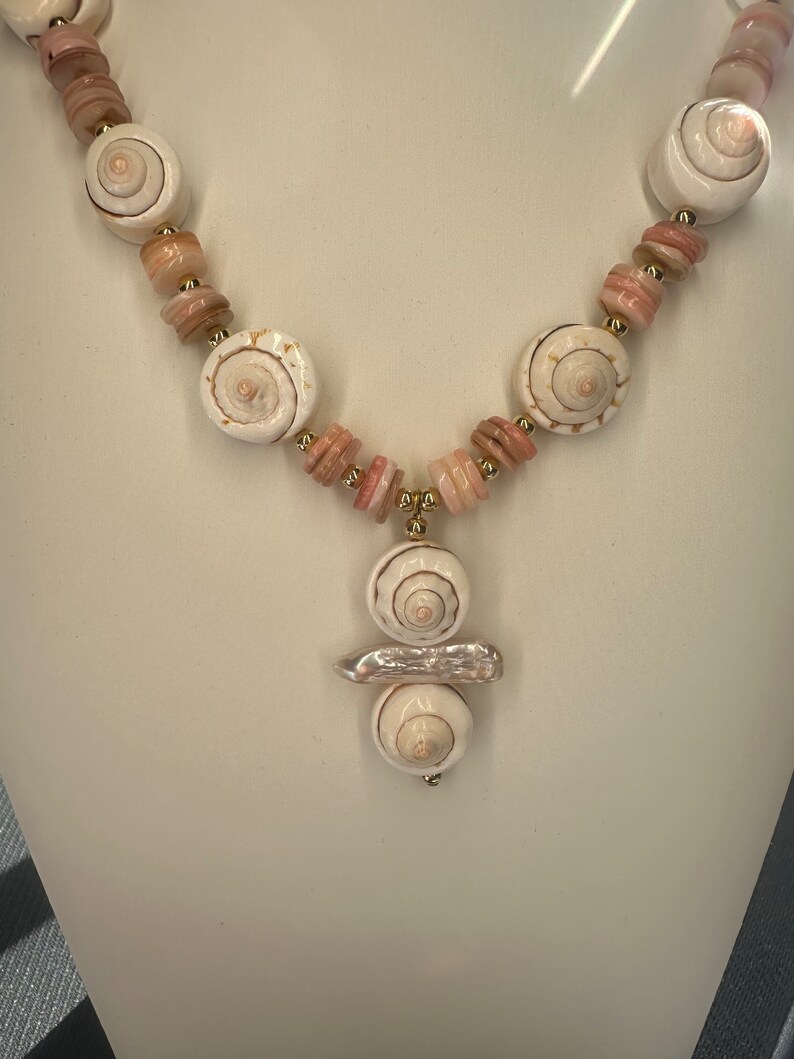 May include: A white and pink shell necklace with a pendant made of three shells. The pendant is attached to the necklace with a gold chain.