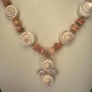May include: A white and pink shell necklace with a gold accent. The necklace features a pendant made of three stacked shells.