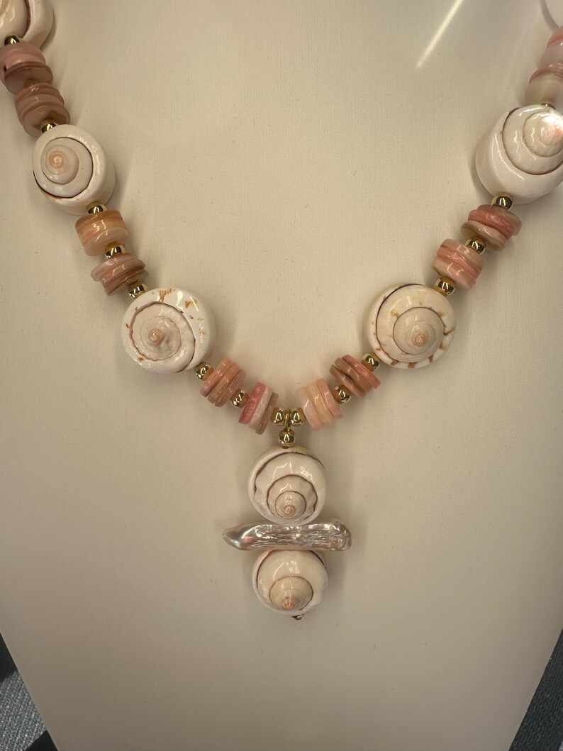 May include: A white and pink shell necklace with a gold accent. The necklace features a pendant made of three stacked shells.