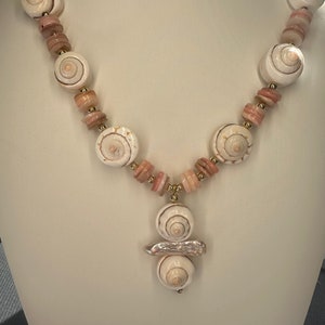 May include: A white and pink shell necklace with a large shell pendant. The necklace is made of natural shells and has a gold accent.