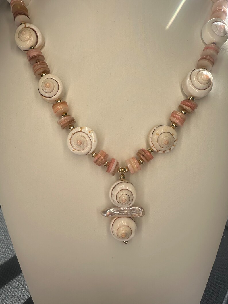 May include: A white and pink shell necklace with a large shell pendant. The necklace is made of natural shells and has a gold accent.