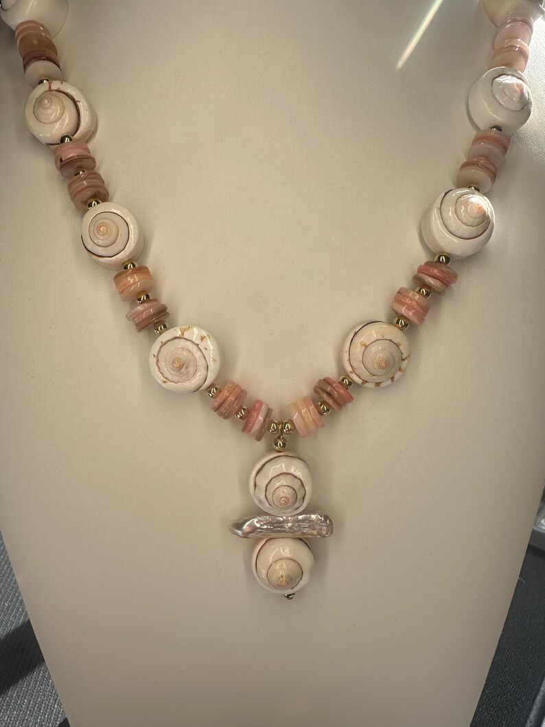 May include: A white seashell necklace with pink and gold beads. The necklace has a pendant with three white seashells.