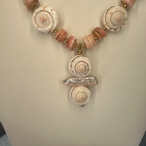 May include: A white and pink shell necklace with a pendant featuring three stacked shells. The pendant is attached to the necklace with a gold chain.