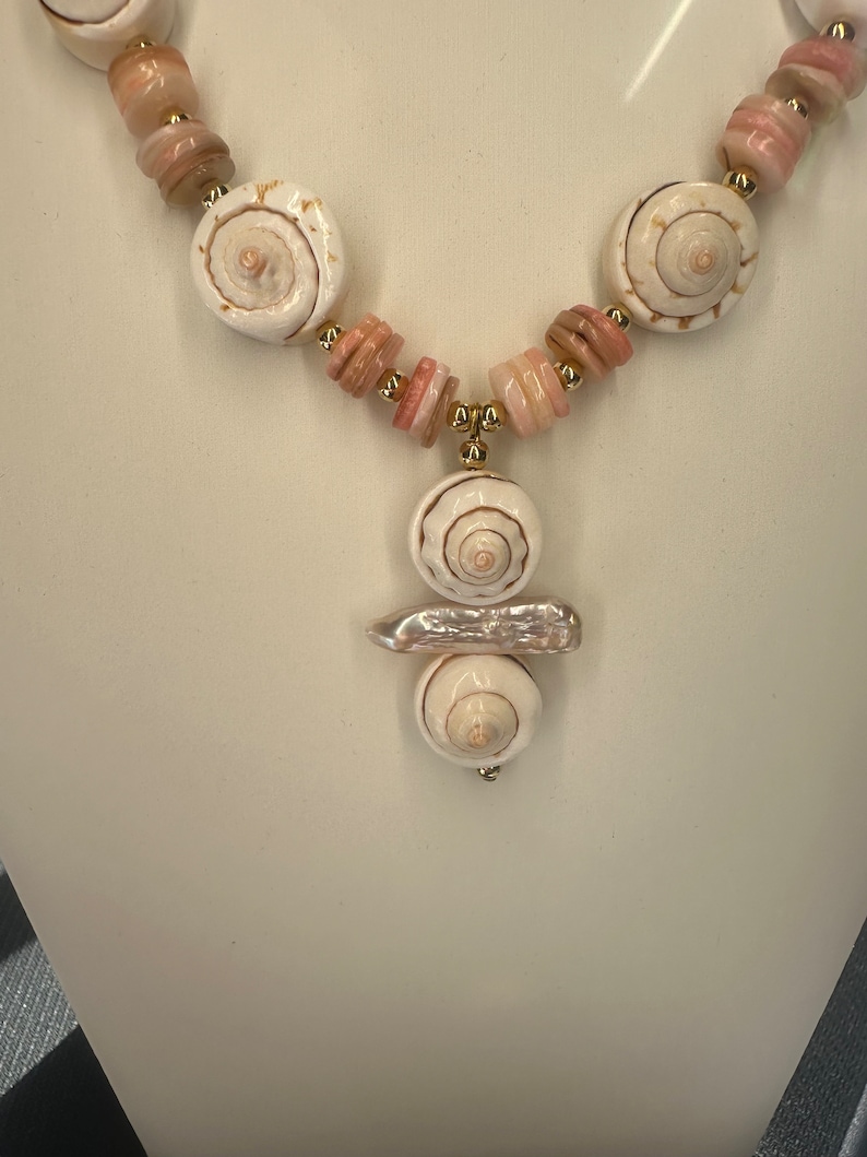 May include: A white and pink shell necklace with a pendant featuring three stacked shells. The pendant is attached to the necklace with a gold chain.