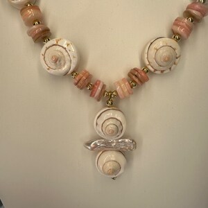 May include: A white and pink shell necklace with a gold accent. The necklace features a pendant with three stacked shells.