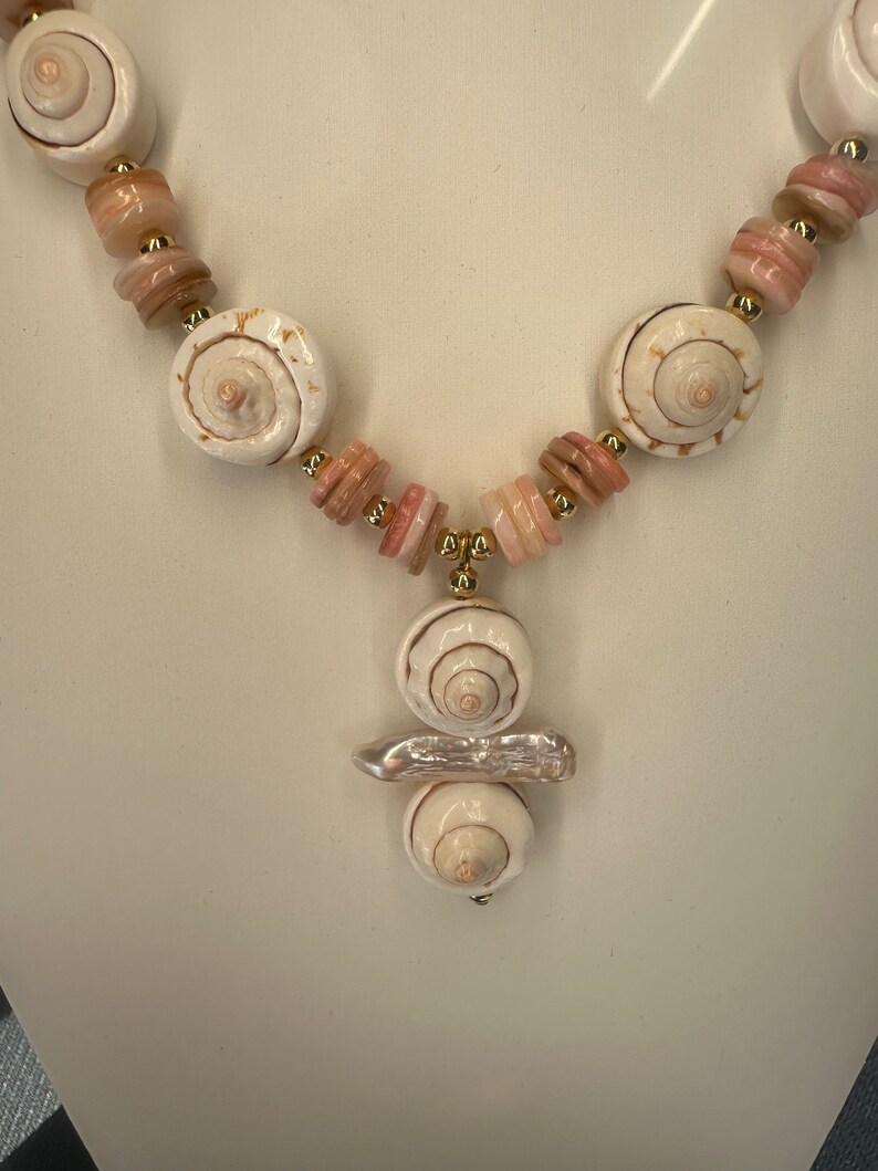 May include: A white and pink shell necklace with a gold accent. The necklace features a pendant with three stacked shells.