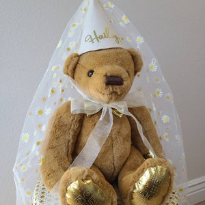 May include: A brown teddy bear wearing a white tulle veil with gold daisy accents and a white felt hat with the name "Hailey" written in gold lettering. The bear is sitting on a yellow wicker chair.