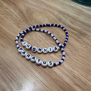 May include: Two beaded bracelets with white beads and blue accents. The bracelets spell out the words "INOMNIA" and "PARATUS".