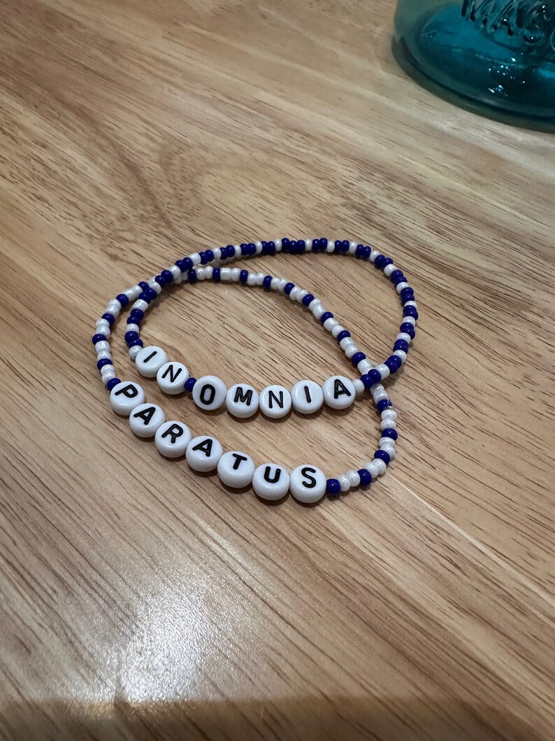 May include: Two beaded bracelets with white beads and blue accents. The bracelets spell out the words "INOMNIA" and "PARATUS".