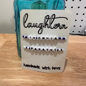 May include: A white card with black text that reads "laughlorn" and "handmade with love". Two beaded bracelets are attached to the card, one says "INOMNIA" and the other says "PARATUS".