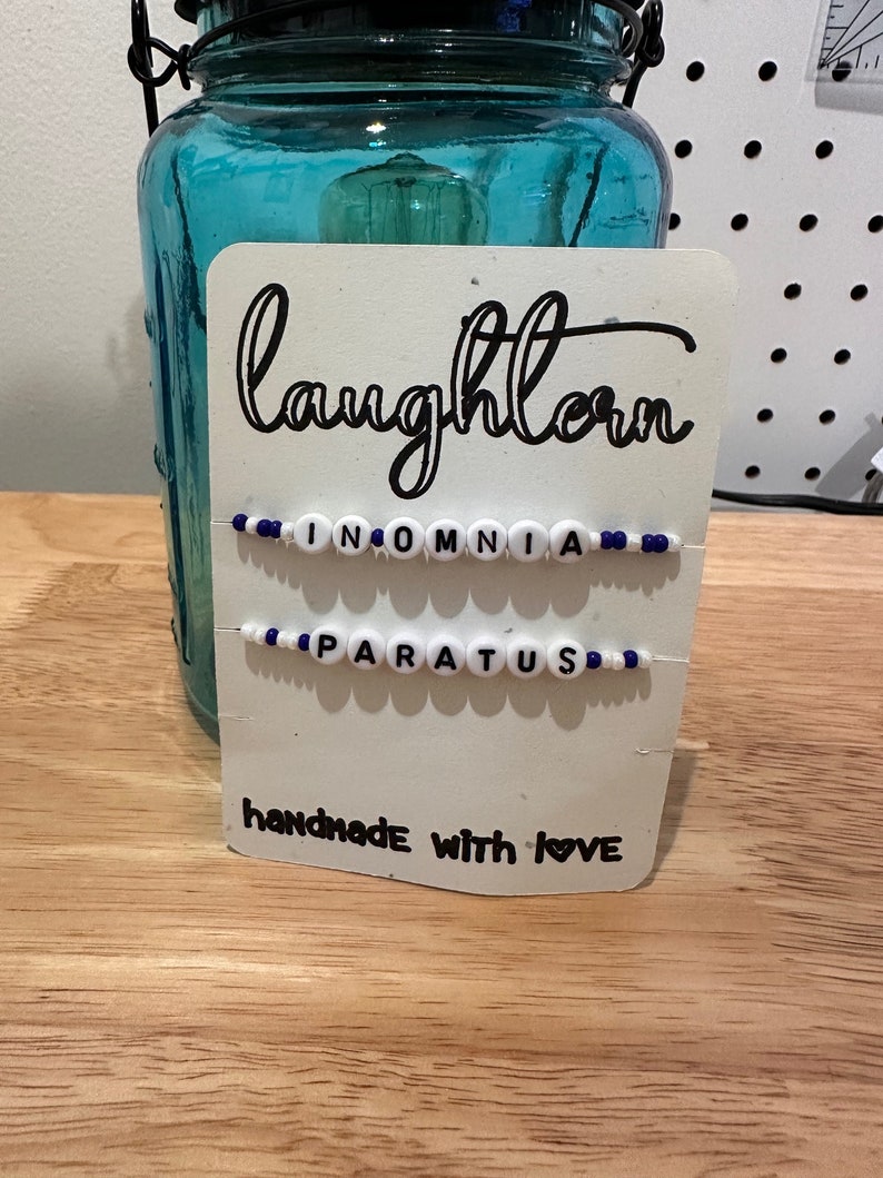 May include: A white card with black text that reads "laughlorn" and "handmade with love". Two beaded bracelets are attached to the card, one says "INOMNIA" and the other says "PARATUS".
