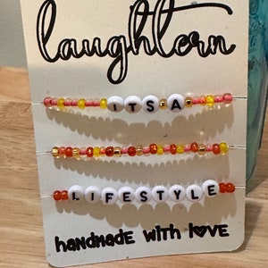 May include: White card with black text that reads "laughlove" and "handmade with love". Three beaded bracelets with white letter beads spelling "IT'S A", "LIFESTYLE", and "LOVE". The beads are red, orange, yellow, and gold.