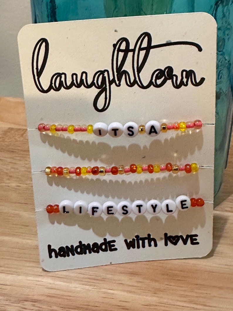 May include: White card with black text that reads "laughlove" and "handmade with love". Three beaded bracelets with white letter beads spelling "IT'S A", "LIFESTYLE", and "LOVE". The beads are red, orange, yellow, and gold.