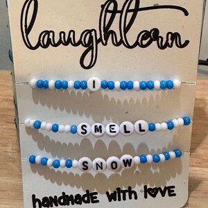 May include: Three beaded bracelets with the words "I Smell Snow" and the text "handmade with love" on a white card with the word "laughlorn" written in black cursive.