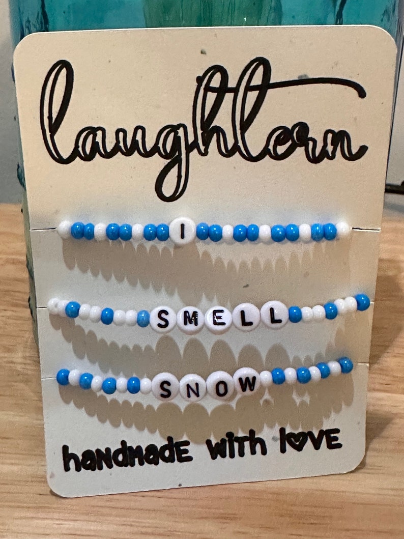 May include: Three beaded bracelets with the words "I Smell Snow" and the text "handmade with love" on a white card with the word "laughlorn" written in black cursive.