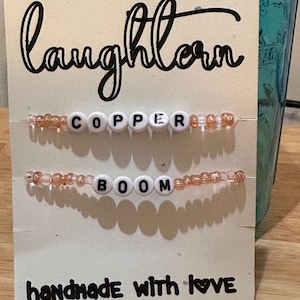 May include: A white card with the word "laughlove" written in black cursive. The card has two beaded bracelets on it, one that says "COPPER" and one that says "BOOM". The card also says "handmade with love" in black cursive.