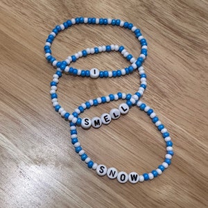 May include: Three beaded bracelets with alternating blue and white beads. The bracelets spell out "I", "SMELL", and "SNOW" in white letters.