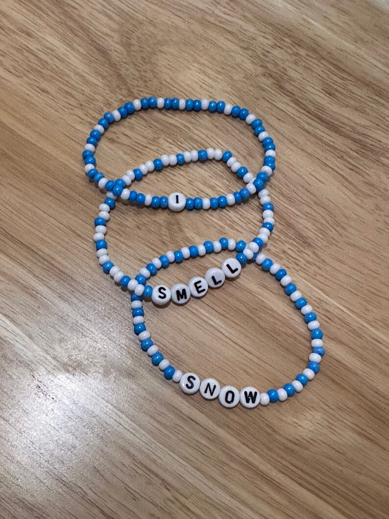 May include: Three beaded bracelets with alternating blue and white beads. The bracelets spell out "I", "SMELL", and "SNOW" in white letters.