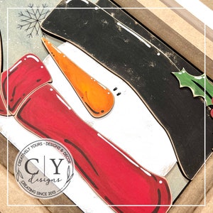 May include: A wooden snowman craft kit with a red hat, black top hat, white body, orange nose, and black eyes. The kit includes a holly sprig with red berries. The kit is made by "CY Designs" and says "Creating Since 2013".
