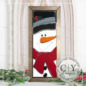 May include: A framed wooden sign with a painted snowman wearing a black hat, gray scarf, and red scarf. The snowman has a carrot nose, black eyes, and a black mouth. The sign is decorated with a green holly sprig with red berries.