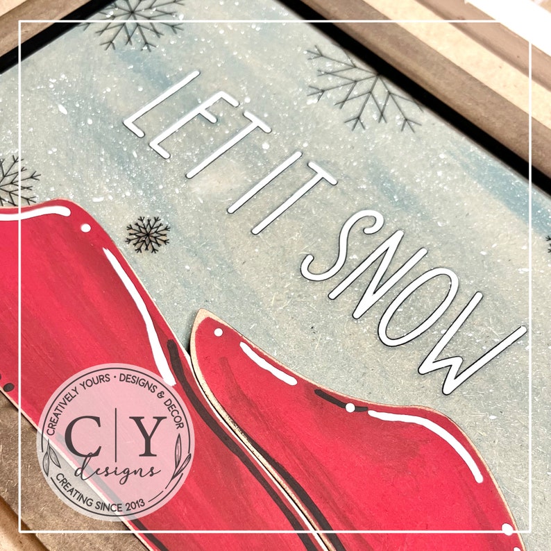 May include: Red wooden cutout of a pair of boots with white outlines against a blue and white background with the text "Let it snow" in white. The image includes a small logo in the bottom left corner that reads "Creatively Yours - Designs & Decor CY Designs Creating Since 2013."