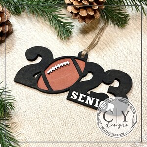 May include: A black wooden ornament shaped like the number 2022 with a brown football in the centre. The ornament also has the word "SENI" written on it. The ornament is decorated with a white and brown design. The ornament is hanging from a string. The ornament is made by "CY Designs" and says "CREATIVELY YOURS - DESIGNS & DECOR CREATING SINCE 2013".