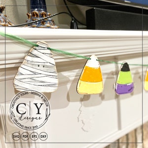 Puede incluir: Un bander&iacute;n de Halloween con recortes de madera de una momie, ma&iacute;z dulce y un fantasma. El bander&iacute;n est&aacute; enhebrado con una cinta verde y cuelga de una repisa blanca. El bander&iacute;n incluye el texto "Creatively Yours - Designs & Decor CY Designs Creating Since 2013".