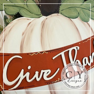 Puede incluir: Una calabaza blanca con detalles de pintura marr&oacute;n y beige, con una pancarta roja que dice "Give Thanks" en escritura blanca. La pancarta tiene un borde con patr&oacute;n dorado y marr&oacute;n. La pancarta tambi&eacute;n tiene un peque&ntilde;o logotipo que dice "Creatively Yours Designs & Decor CY Designs Creating Since 2013".