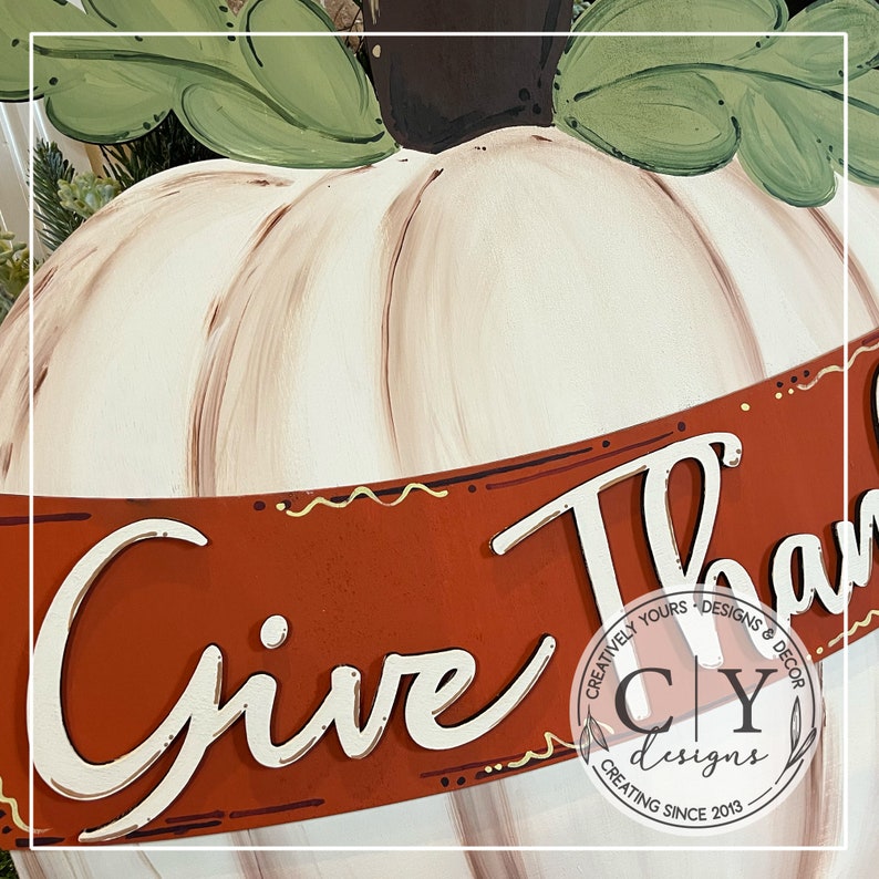 Puede incluir: Una calabaza blanca con detalles de pintura marr&oacute;n y beige, con una pancarta roja que dice "Give Thanks" en escritura blanca. La pancarta tiene un borde con patr&oacute;n dorado y marr&oacute;n. La pancarta tambi&eacute;n tiene un peque&ntilde;o logotipo que dice "Creatively Yours Designs & Decor CY Designs Creating Since 2013".
