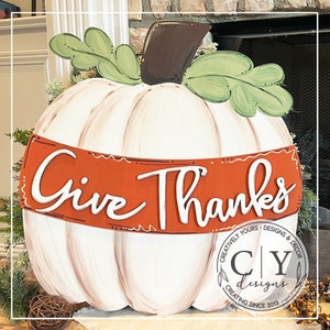 Puede incluir: Calabaza blanca con un tallo marr&oacute;n y hojas verdes. La calabaza tiene una pancarta roja con las palabras "Give Thanks" en letras blancas. La calabaza est&aacute; decorada con un dise&ntilde;o de pintura marr&oacute;n y blanco. La parte inferior de la calabaza tiene un logotipo marr&oacute;n y blanco con el texto "Creatively Yours - Designs & Decor CY Designs Creating Since 2013."