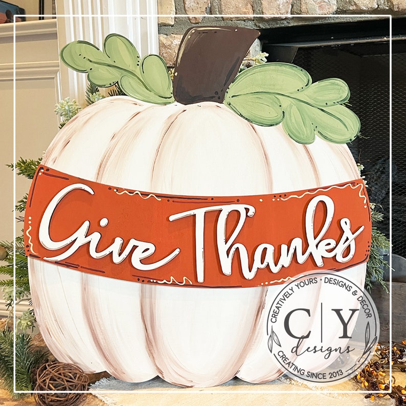 Puede incluir: Calabaza blanca con un tallo marr&oacute;n y hojas verdes. La calabaza tiene una pancarta roja con las palabras "Give Thanks" en letras blancas. La calabaza est&aacute; decorada con un dise&ntilde;o de pintura marr&oacute;n y blanco. La parte inferior de la calabaza tiene un logotipo marr&oacute;n y blanco con el texto "Creatively Yours - Designs & Decor CY Designs Creating Since 2013."