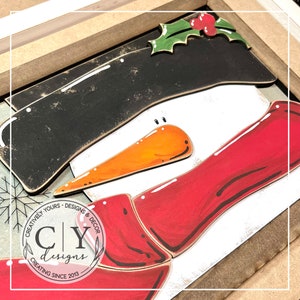 May include: A wooden snowman craft project with a black hat, orange carrot nose, and red scarf. The snowman is partially assembled and has a white background. The image includes the text "Creatively Yours - Designs & Decor CY Designs Creating Since 2013".