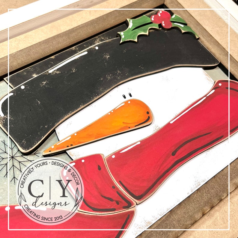 May include: A wooden snowman craft project with a black hat, orange carrot nose, and red scarf. The snowman is partially assembled and has a white background. The image includes the text "Creatively Yours - Designs & Decor CY Designs Creating Since 2013".