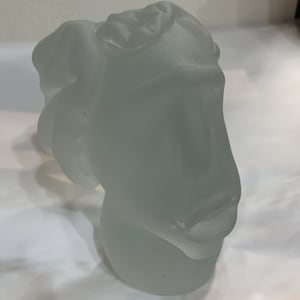 Könnte beinhalten: Eine frostige Glasskulptur eines Gesichts mit einem stilisierten, abstrakten Design. Die Skulptur ist weiß und hat eine glatte, matte Oberfläche.