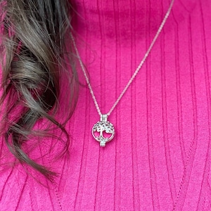 May include: A silver necklace with a tree of life pendant. The necklace is worn by a person wearing a pink sweater.