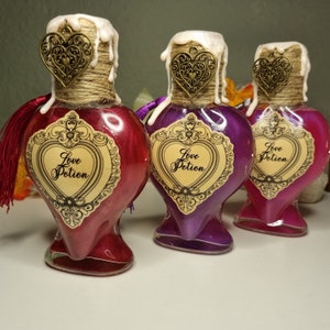 Könnte beinhalten: Drei herzförmige Glasflaschen, gefüllt mit schimmernder roter, lila und rosa Flüssigkeit. Jede Flasche hat ein dekoratives Etikett mit der Aufschrift "Love Potion". Die Flaschen sind mit einem herzförmigen Anhänger, Bindfaden und einer wachsartigen Substanz versehen.