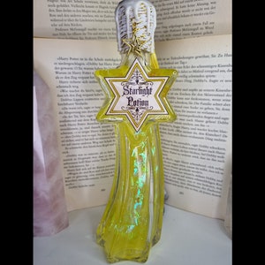 Könnte beinhalten: Eine gelbe Glasflasche mit einem sternförmigen Etikett mit der Aufschrift "Starlight Potion". Die Flasche hat eine einzigartige Form und ist mit einer schimmernden, irisierenden Flüssigkeit gefüllt. Die Flasche ist mit einem weißen und goldenen Verschluss versehen.