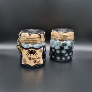 Könnte beinhalten: Zwei Gläser gefüllt mit schwarzen und blauen Kugeln, beschriftet mit "Aragog's Eggs". Jedes Glas hat einen schwarzen Wachsdeckel, Bindfaden und einen Anhänger. Ein Glas hat einen Spinnenanhänger, das andere einen Heiligtümer des Todes-Anhänger. Auf schwarzem Hintergrund.