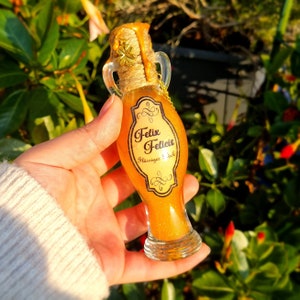 Puede incluir: Una botella de vidrio decorativa llena de líquido dorado brillante, etiquetada como "Felix Felicis". La botella tiene una tapa de corcho adornada con un amuleto de trébol de cuatro hojas dorado y está envuelta con hilo.