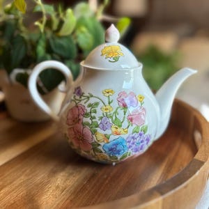May include: A white ceramic teapot featuring a colorful hand-painted floral pattern with pink, blue, yellow, and purple blossoms. The teapot has a curved handle, a matching lid with a small finial, and a delicate spout, all decorated with flowers.