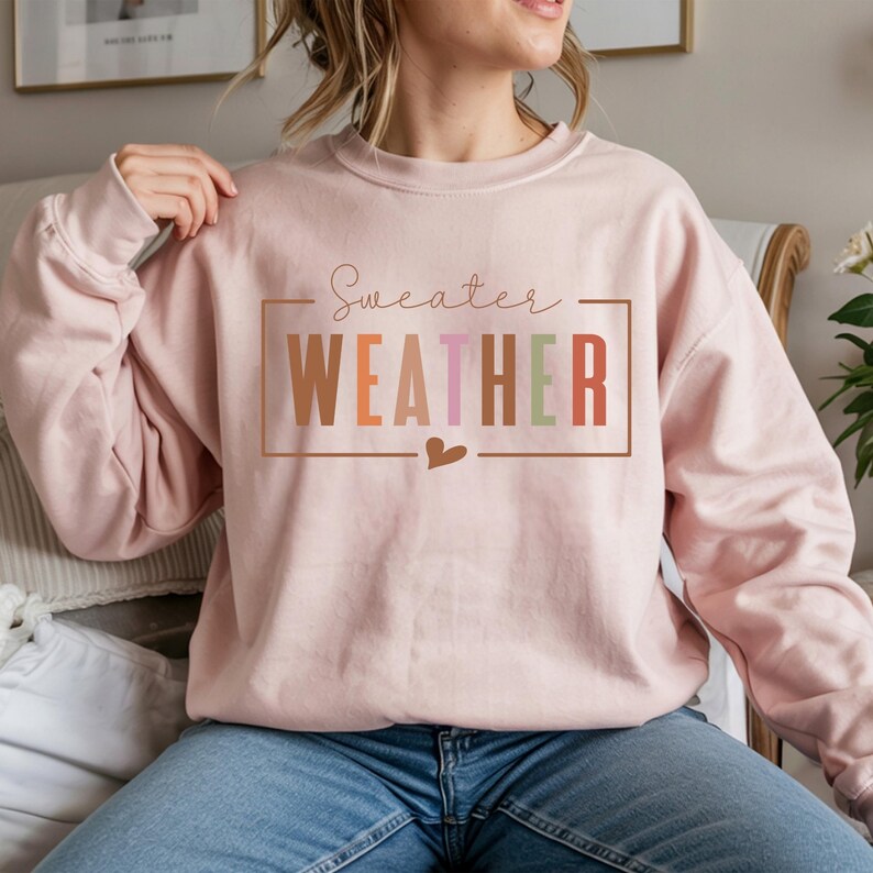 Puede incluir: Una sudadera rosa claro con la frase "Sweater Weather" en un marco rectangular. La palabra "WEATHER" est&aacute; en letras de bloque coloridas. Un peque&ntilde;o coraz&oacute;n est&aacute; debajo del marco. La sudadera es de estilo casual.