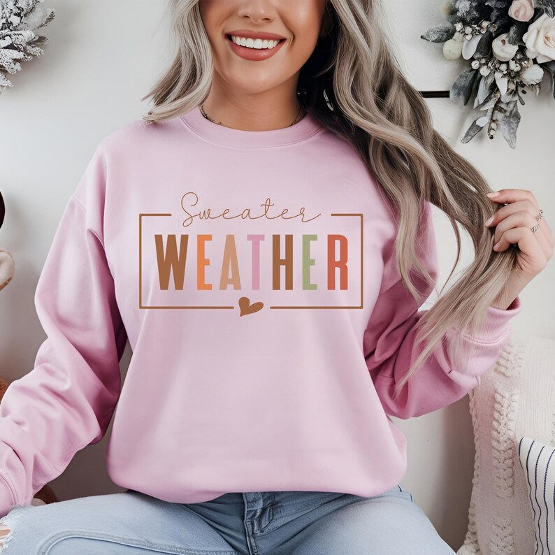 Puede incluir: Sudadera rosa claro con la frase "Sweater Weather" en un marco rectangular. El texto es de varios colores, incluyendo marr&oacute;n, naranja, rosa, verde y blanco. Un peque&ntilde;o coraz&oacute;n est&aacute; debajo del texto.