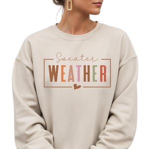 Puede incluir: Sudadera beige con la frase "Sweater Weather" en un marco rectangular. El texto es de varios colores, incluyendo marr&oacute;n, naranja, rosa y verde. Un peque&ntilde;o coraz&oacute;n est&aacute; debajo del texto.