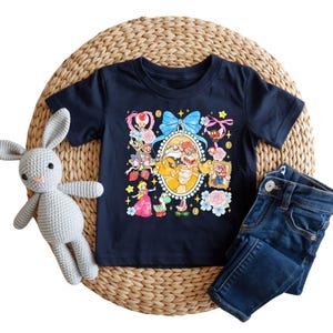 May include: Navy blue t-shirt featuring a colorful graphic of Bowser and other characters from the Super Mario video game series. The shirt is paired with dark wash denim jeans and a gray crocheted bunny.