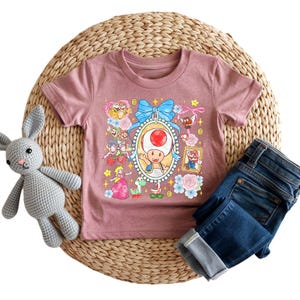 May include: A mauve short-sleeved t-shirt featuring a colorful graphic of a cartoon character in a frame, surrounded by other characters and floral accents. A pair of blue jeans is folded next to the shirt. A gray stuffed bunny is on the left.