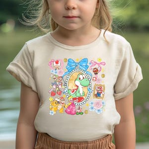 May include: A beige t-shirt featuring a colorful graphic of Yoshi, Mario, Princess Peach, and other characters from the Super Mario franchise. The design includes a blue bow, flowers, and gold coins.