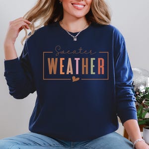 Puede incluir: Sudadera azul marino con la frase "Sweater Weather" en un dise&ntilde;o rectangular. La palabra "WEATHER" est&aacute; en letras de bloque coloridas. Un peque&ntilde;o coraz&oacute;n est&aacute; debajo del texto. La sudadera tiene mangas largas.